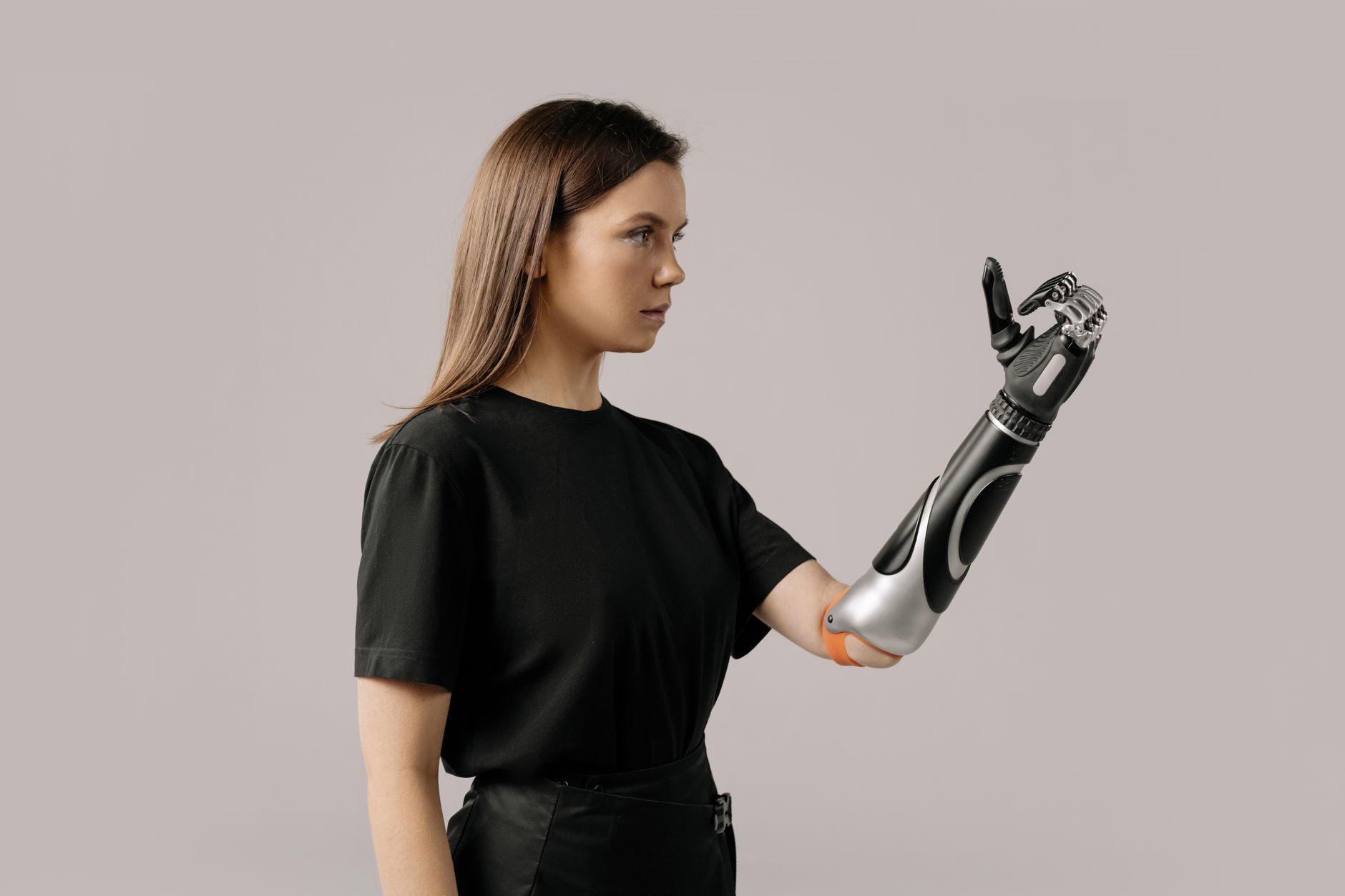 A woman with long hair wearing a black shirt, showing a prosthetic arm while looking thoughtfully at it.