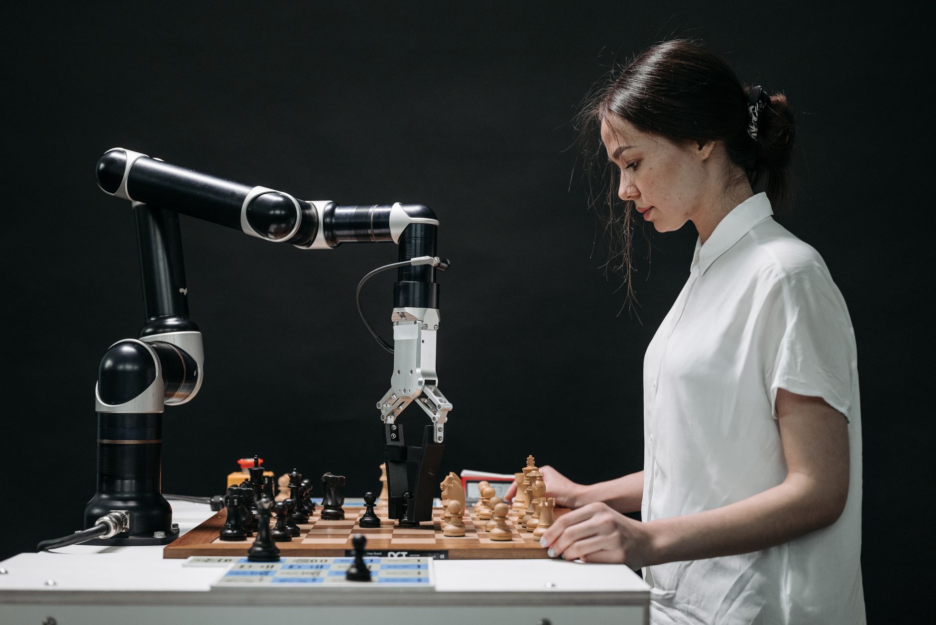 A woman in a white shirt plays chess with a robotic arm beside her, which is reaching towards the chessboard.